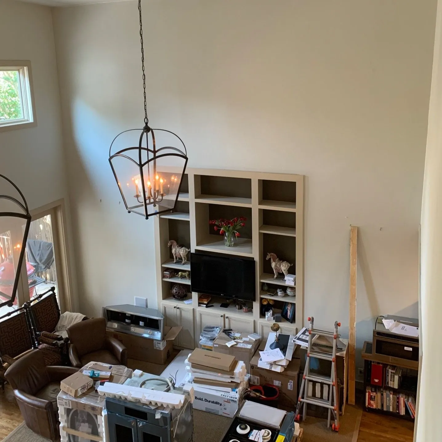 Living room with a large wall-mounted entertainment center, small televisions, decorative items, a ladder, and furniture, with boxes and clutter on the floor.