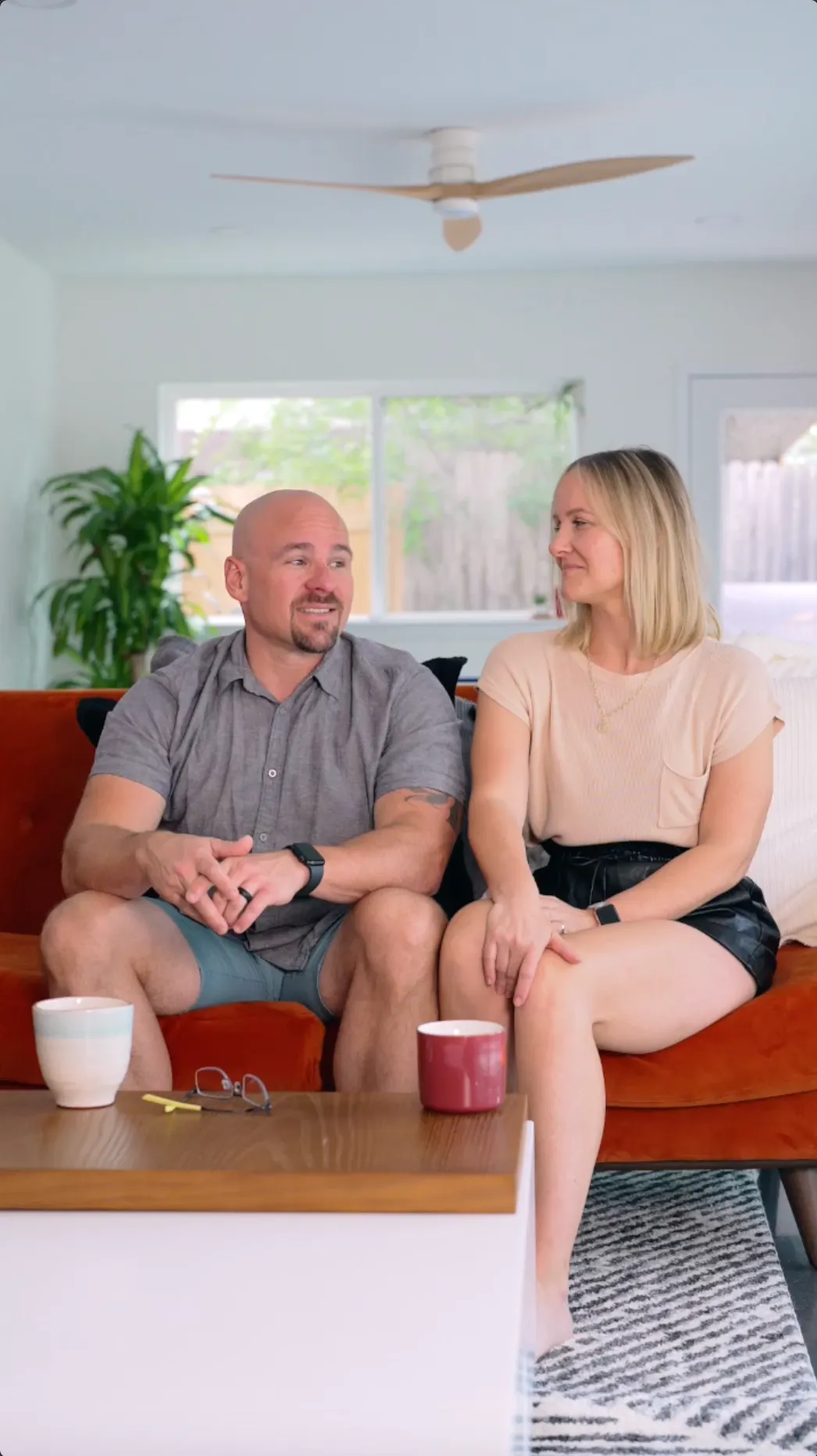 A man and woman sitting on a brown couch in a living room with large windows and houseplants, having a conversation.
