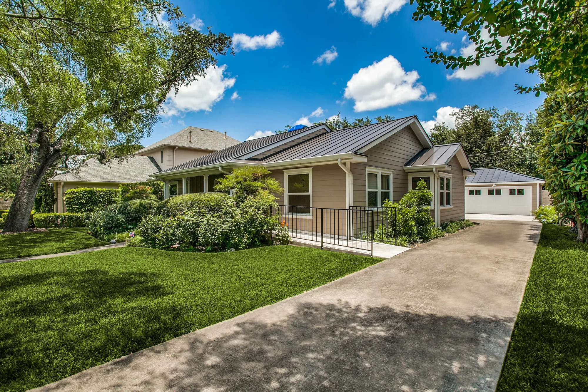 A single-story house with beige siding and a metal roof, connected to a driveway leading to a garage, surrounded by green grass, trees, and shrubs on a sunny day with a partly cloudy sky.