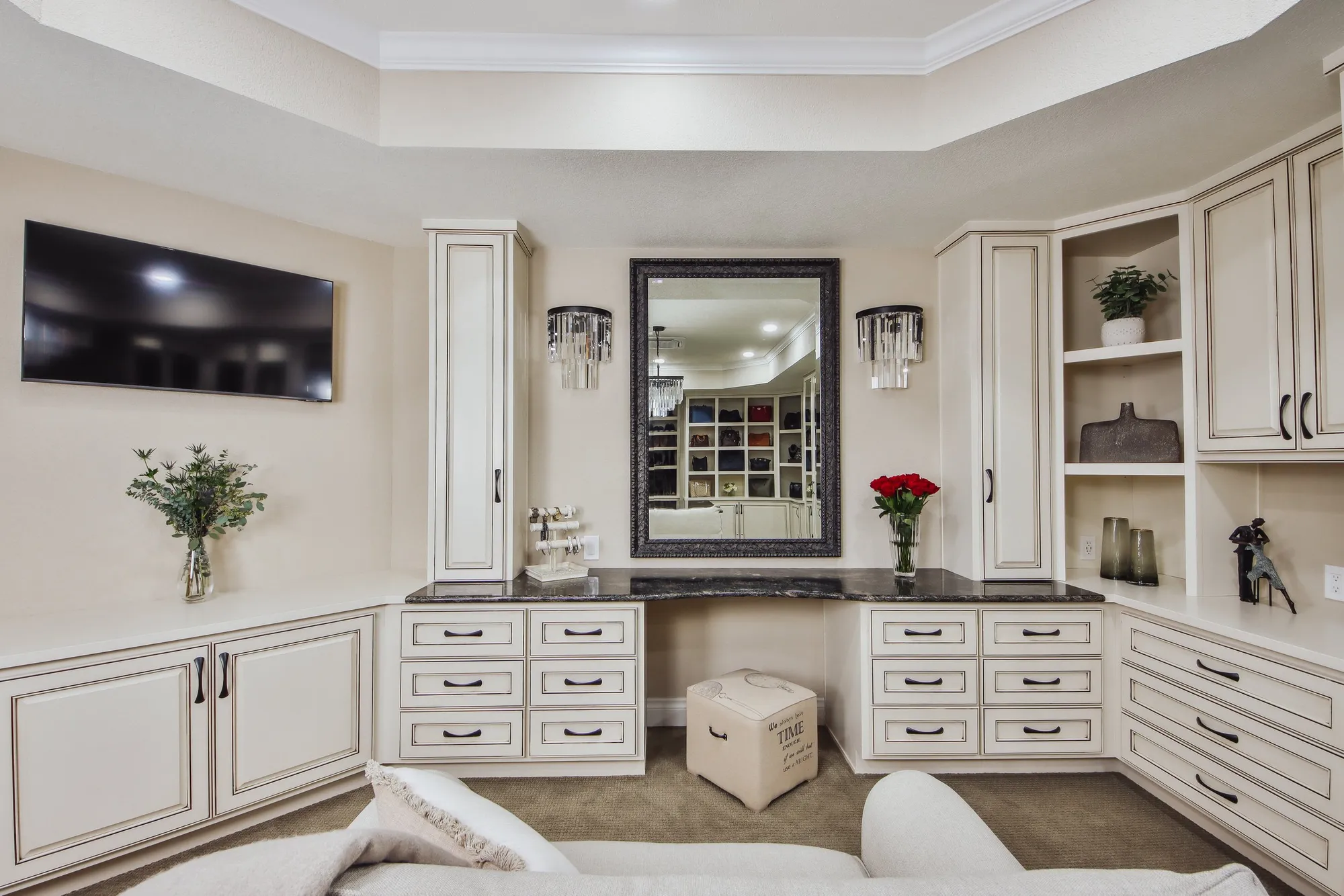 A living room with off-white cabinets and a black marble countertop. A large wall-mounted mirror is centered on the wall, flanked by two modern wall sconces. Decorative items include a vase with red roses, potted plants, and a small animal sculpture. A flat-screen TV is mounted on the left wall, and the reflection in the mirror shows a walk-in closet with shelves of shoes and bags.