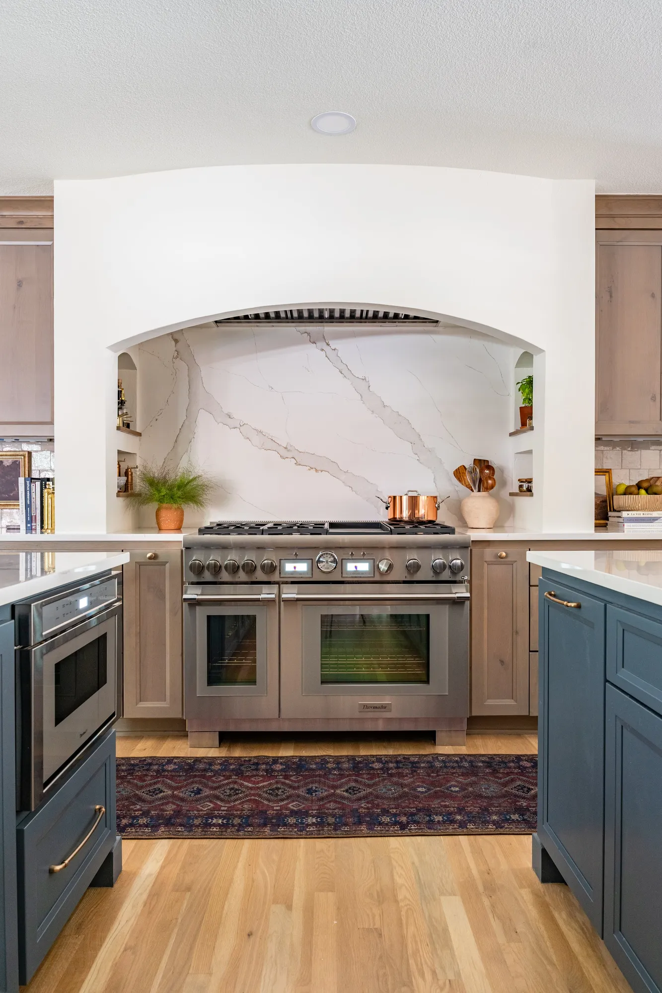 A modern kitchen with a stainless steel oven and stove, blue cabinets on the right, light wood cabinets on each side, and a decorative backsplash with a white marble pattern and gold veining. There is a small shelving nook on each side of the backsplash with potted plants and kitchen items. A marble-patterned archway above the stove. A red and blue patterned rug on a light hardwood floor.