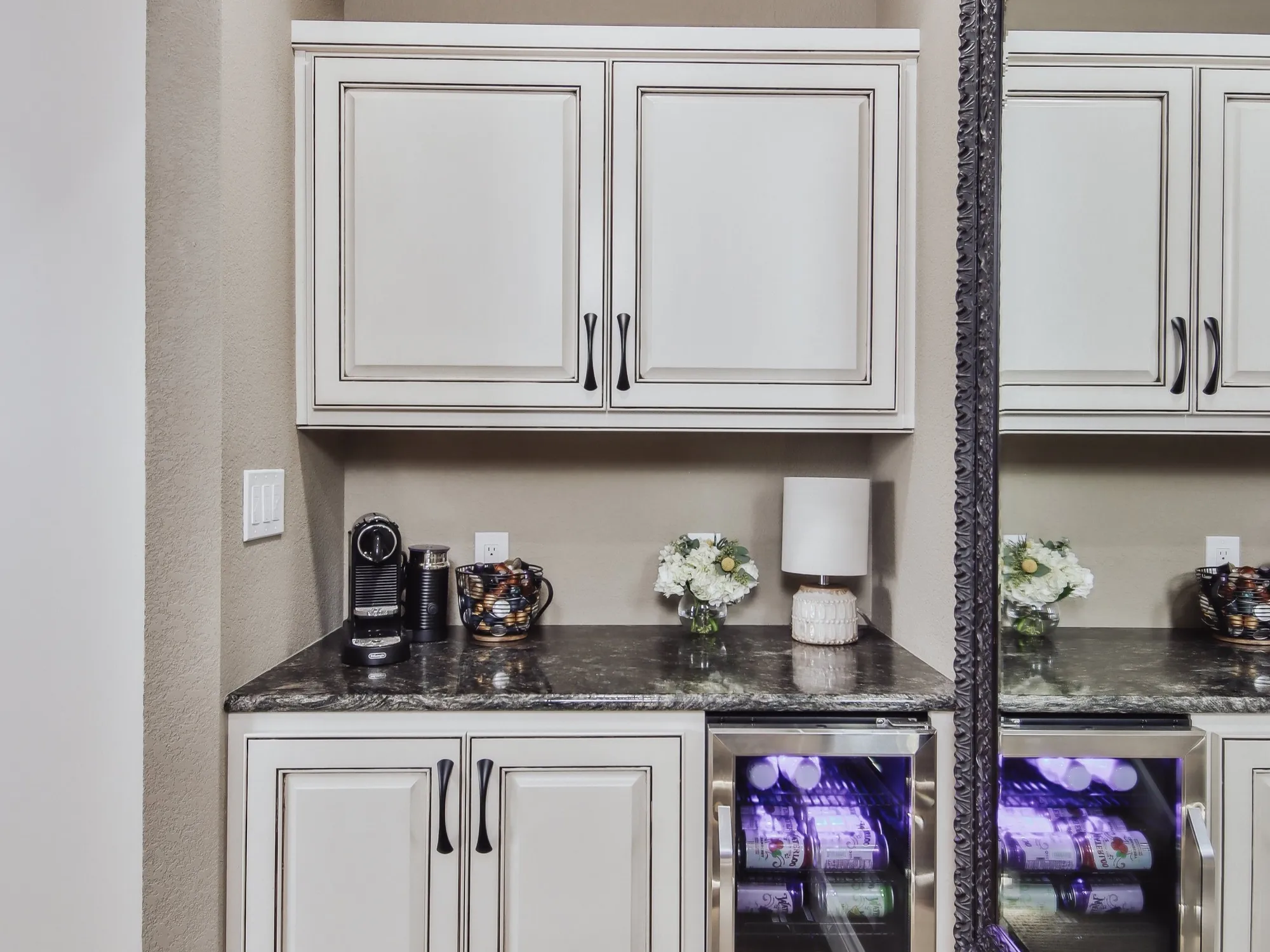 Kitchen countertop with coffee maker, a jar of coffee pods, a flower arrangement, a small lamp, and a mini fridge with beverages, mirror on the right side