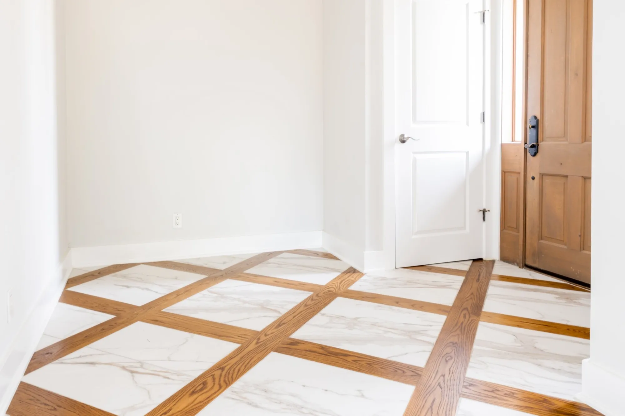 An empty room with white walls, a marble-like tile floor with wood trim, a white door, and a wooden front door.