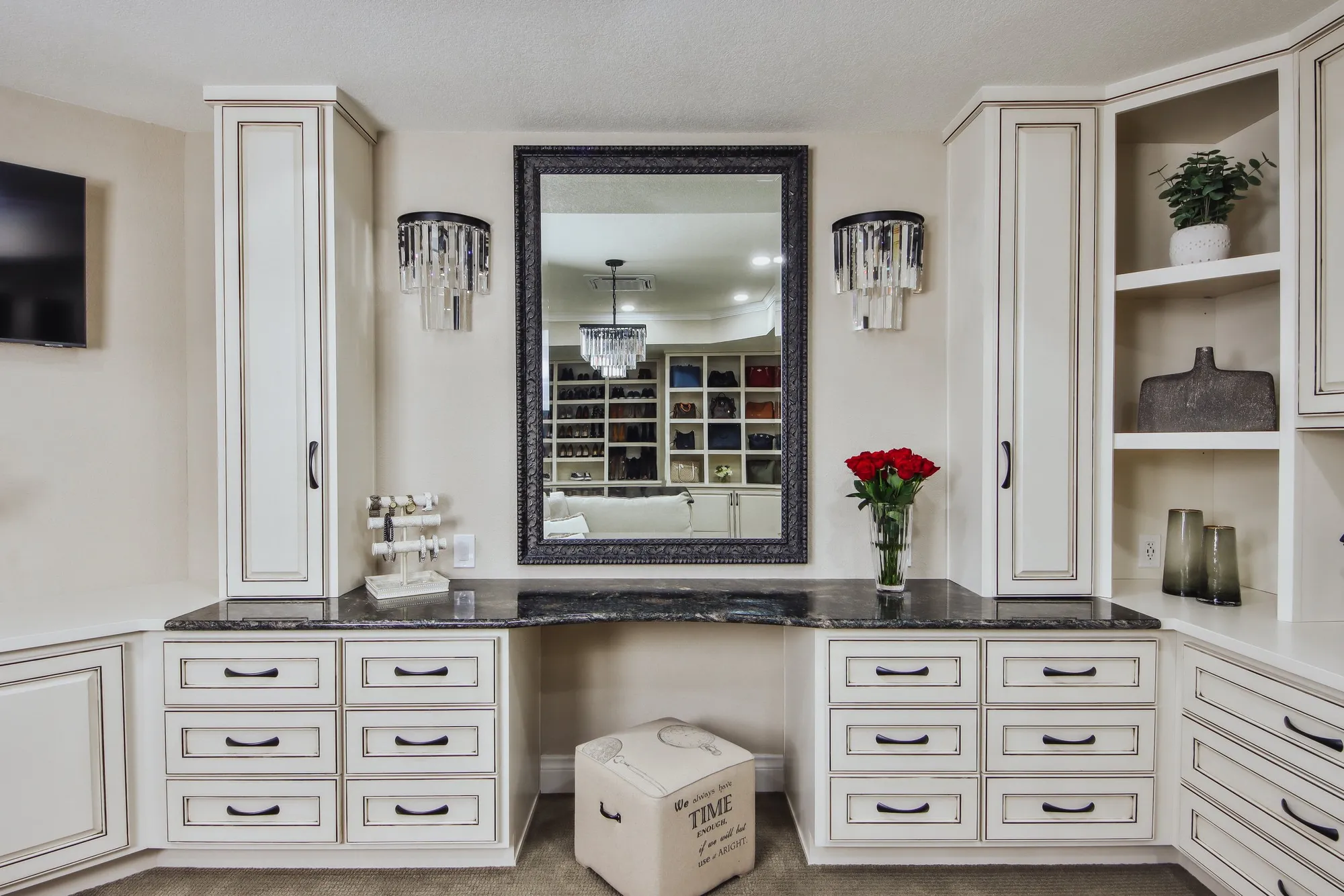 A home bar or vanity area with white cabinetry, black marble countertops, a large framed mirror, and decorative lighting fixtures. There is a vase with red roses on the counter, open shelving with plants and decorative items, and a small upholstered storage box with script writing underneath the mirror.