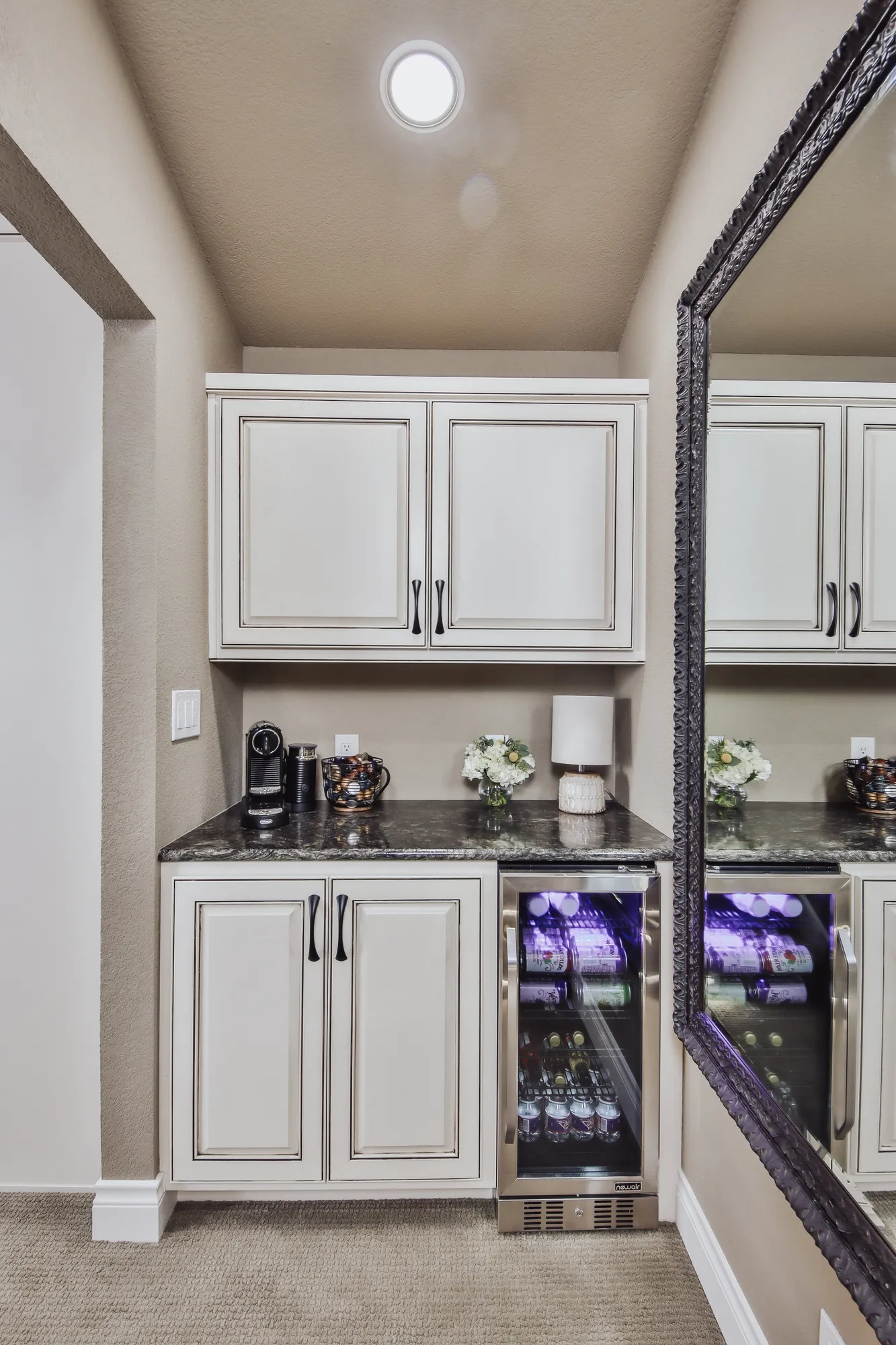A kitchenette area with white cabinets, a dark granite countertop, a small wine refrigerator, and a large wall mirror reflecting the plumbing and decor.