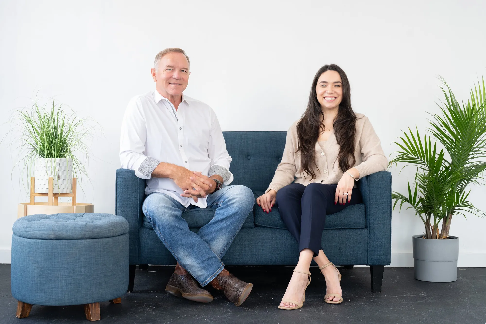 A man and a woman sitting on a blue sofa, smiling at the camera, with green plants on either side and a white wall background.
