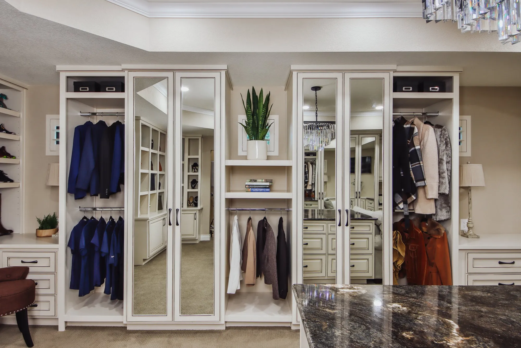 A luxurious bedroom closet with white wood cabinetry, mirrored doors, a potted plant on a small shelf, hanging clothes including suits, jackets, and dresses, and a granite countertop in the foreground.