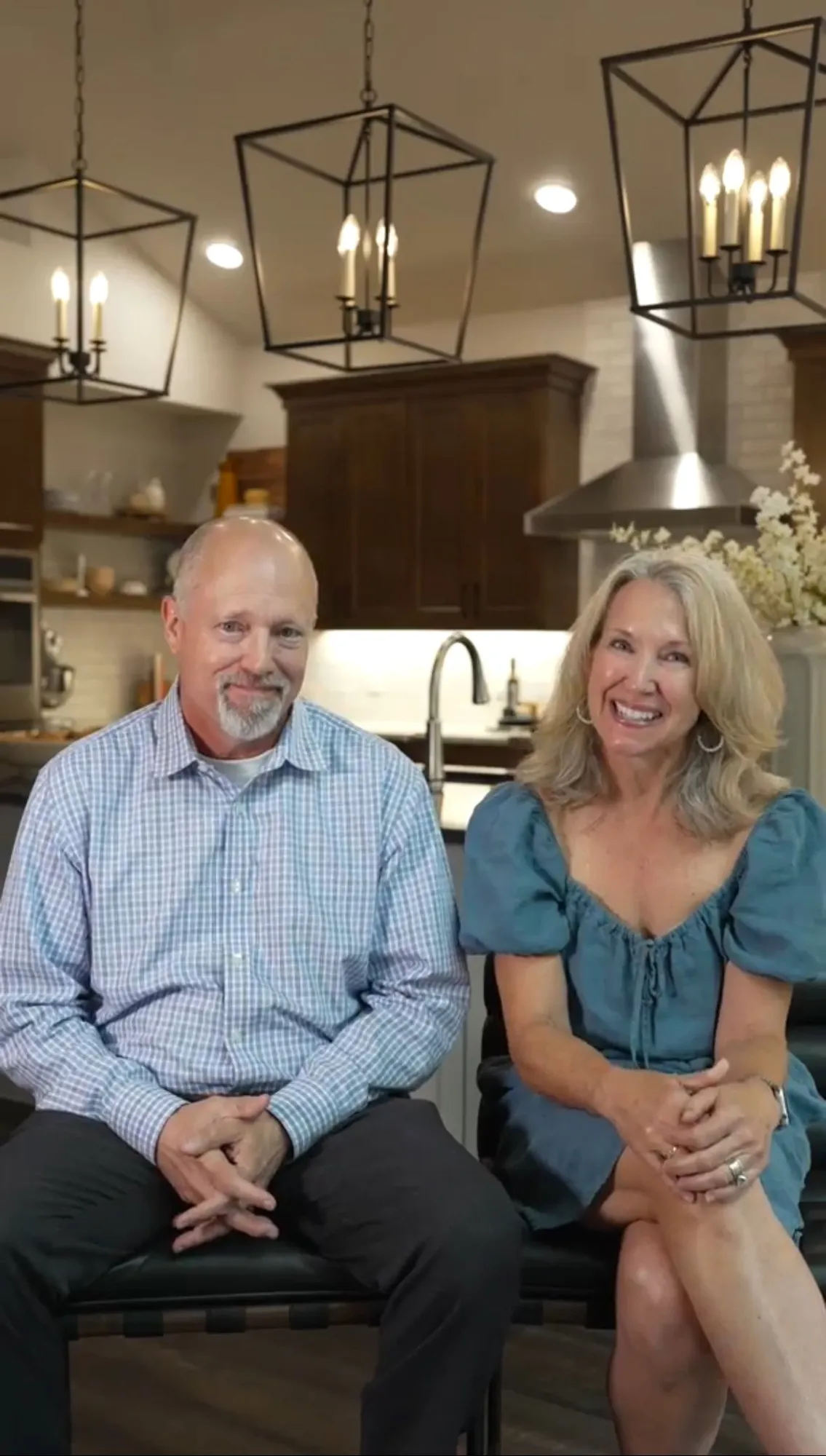 A smiling middle-aged man with a beard and a woman with blonde hair sitting in a modern kitchen with dark wooden cabinets, pendant lights, and a white brick wall in the background.