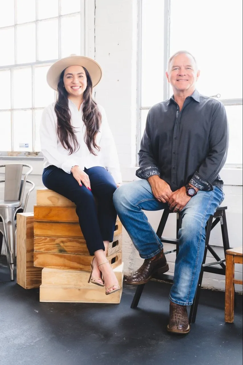 Two people, a young woman and an older man, smiling and sitting indoors near large windows. The woman is sitting on wooden boxes, wearing a white blouse, navy pants, beige heels, and a wide-brimmed hat. The man is sitting on a black stool, wearing a dark button-up shirt, blue jeans, and brown boots.