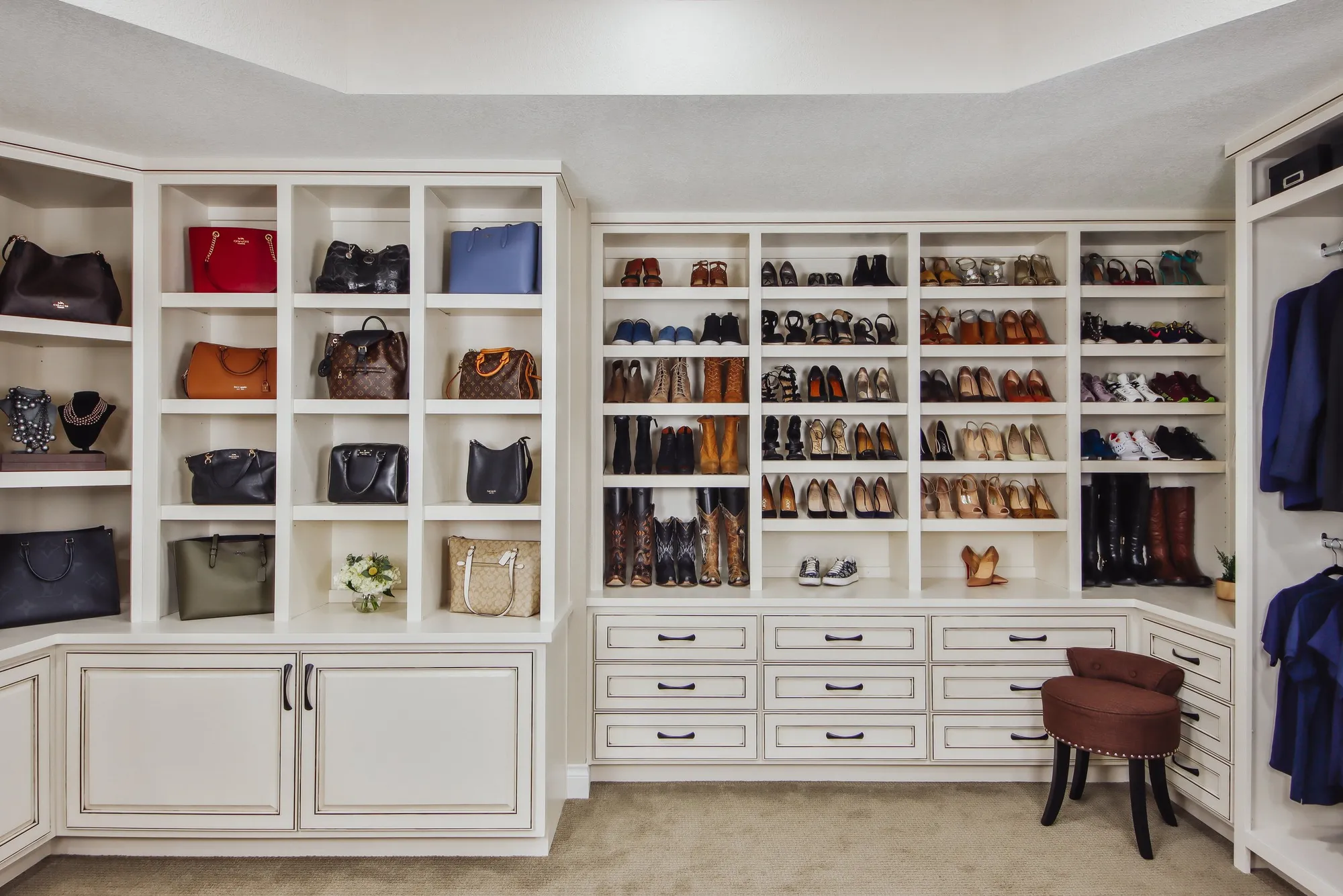 Closet with shelves filled with handbags and shoes, with itt. a small brown ottoman on the right.