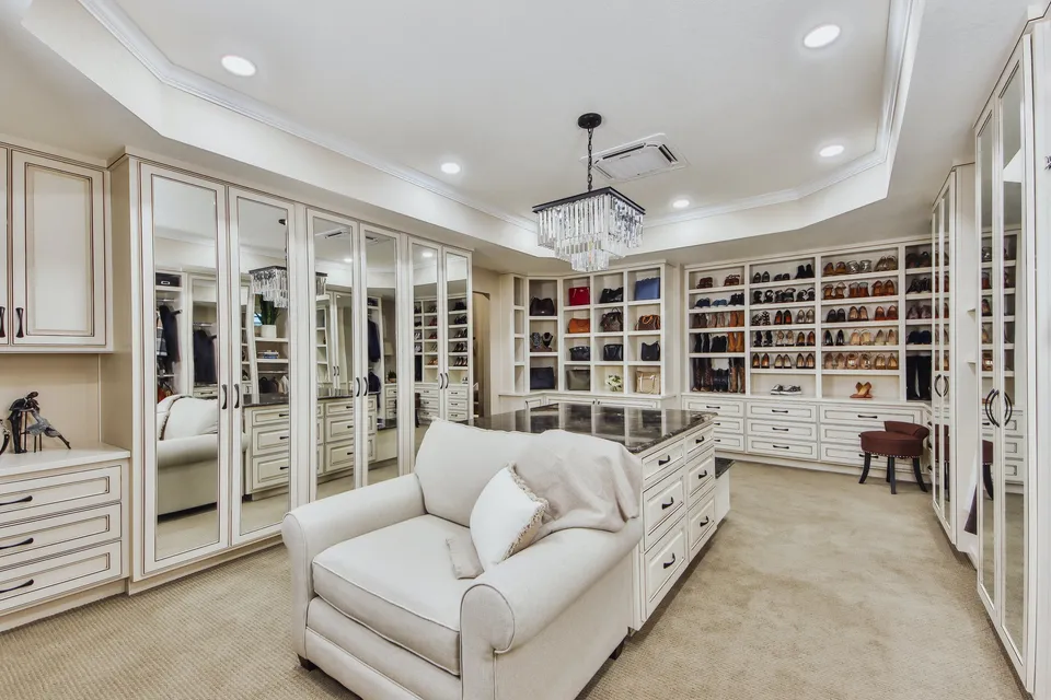 A luxurious walk-in closet with built-in white cabinets, mirrored doors, and display shelving filled with shoes, handbags, and accessories. There is a central island with a dark marble top, a small seating area with a brown upholstered stool, and a white armchair with decorative pillows. A chandelier hangs from the ceiling, and the room is well-lit with recessed lighting.