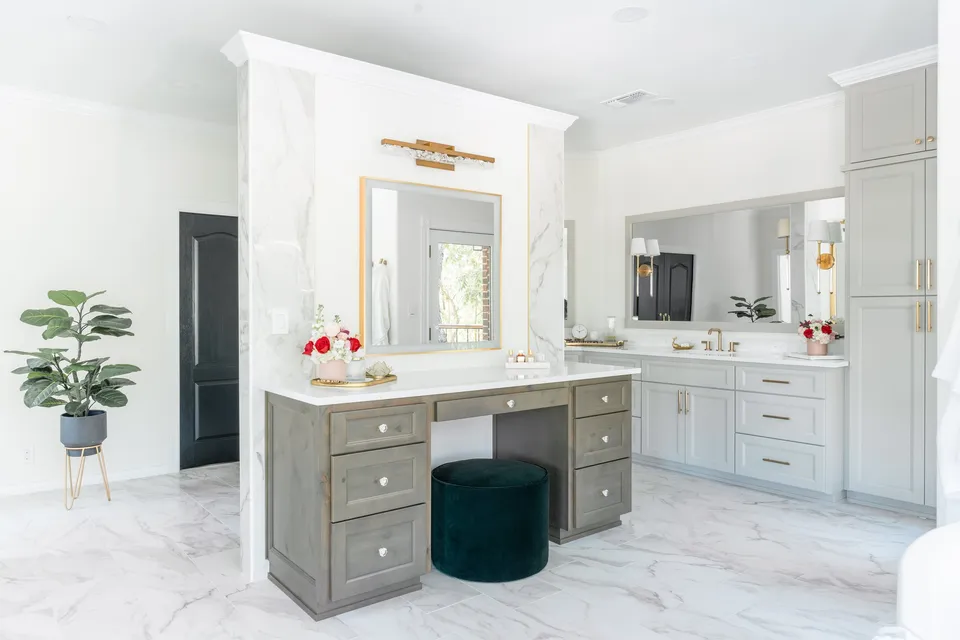 Bright bathroom with white walls and marble flooring, featuring a vanity with a large mirror, a makeup desk with a green velvet stool, and floral arrangements on the counters. There are gold fixtures, a potted plant, and a window visible in the reflection.
