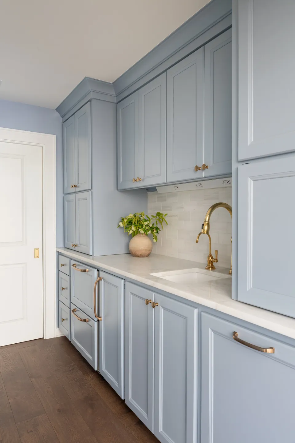 Pale blue kitchen cabinetry with a brass faucet, marble counter and a bowl of greenery