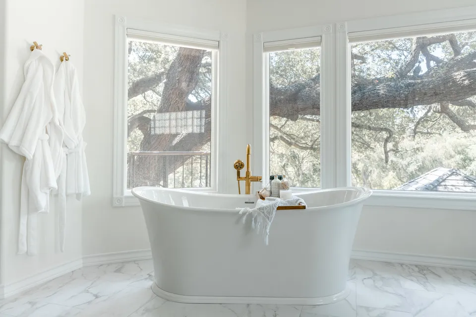 A white bathtub with a gold faucet is in front of large windows showing a view of trees outside. There are white robes hanging on the wall to the left.