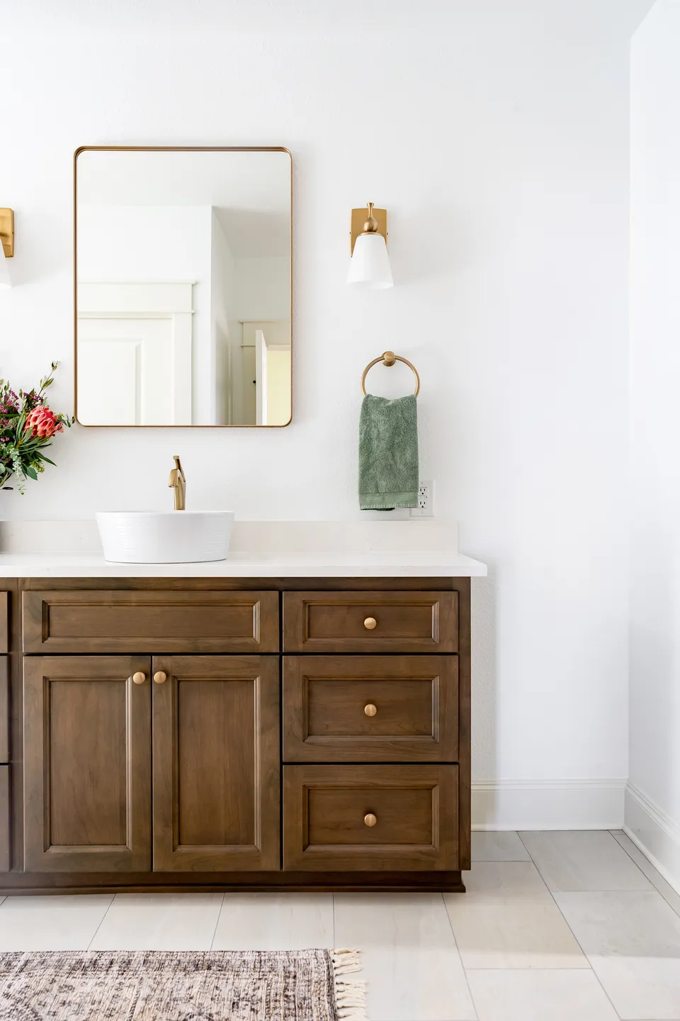 Bathroom vanity with wooden cabinets, a white vessel sink, a gold faucet, a rectangular mirror, wall-mounted light fixtures, a green hand towel on a ring, a floral arrangement on the side, and a small rug on the floor.