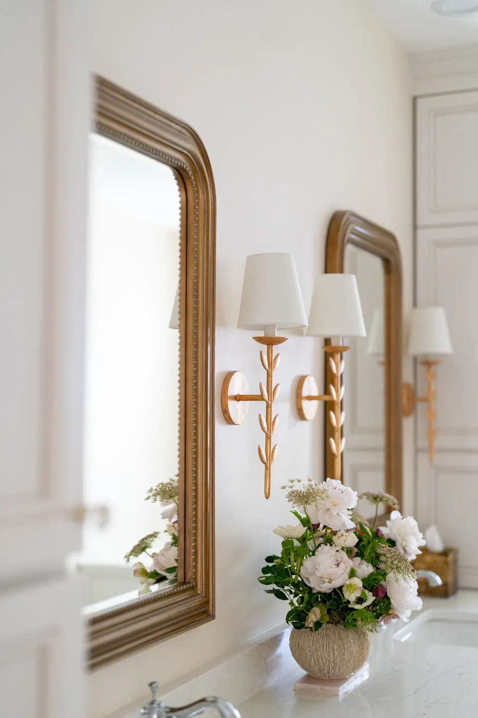 Gilded arched mirrors and brass sconces above a vanity with white flowers