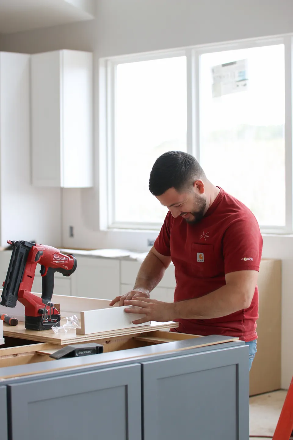 A man in a red shirt working on a woodworking project in a bright, modern kitchen or workshop.