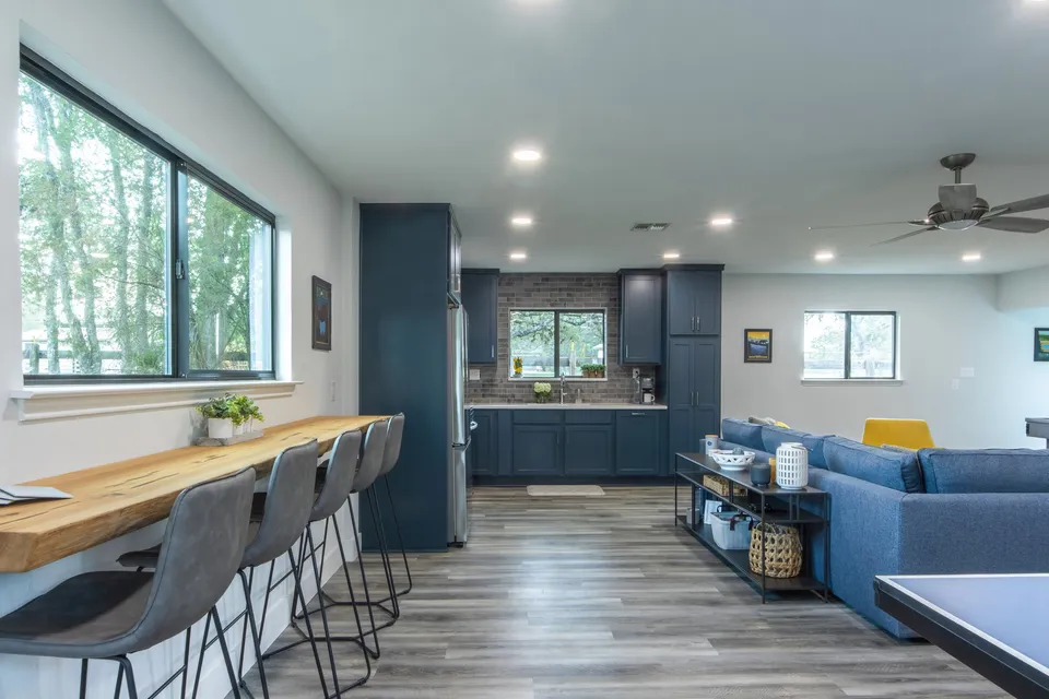 Modern open-concept living room and kitchen with blue cabinets, gray walls, wooden floor, and large windows showing a view of trees outside.