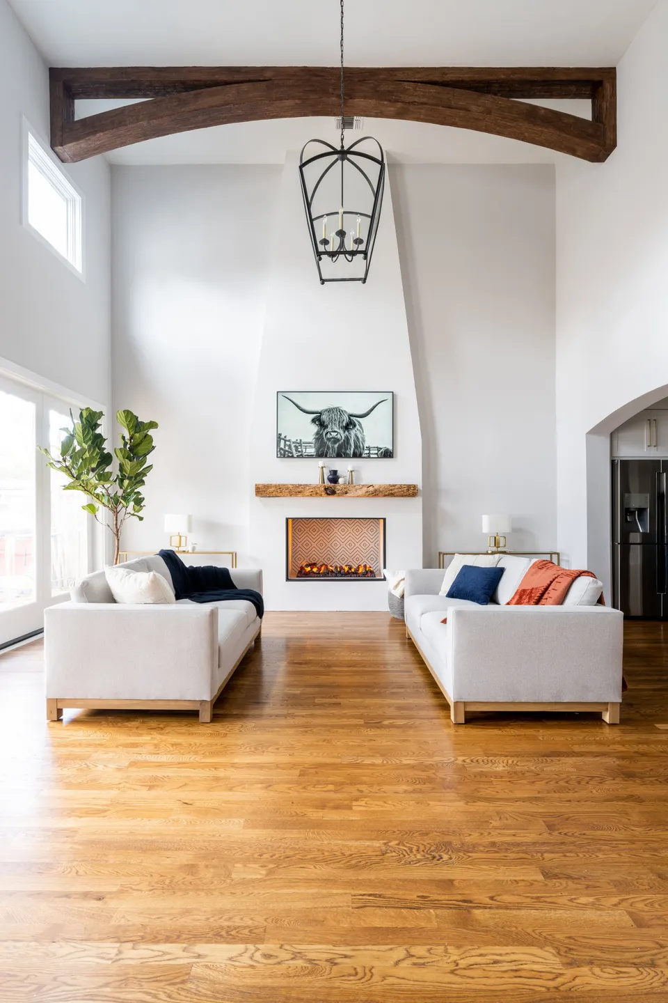 Bright living room with vaulted ceiling, wooden beam, two white sofas with pillows, a fireplace with a wood mantel, a wall-mounted black-and-white portrait of a longhorn steer, and a large window with natural light.